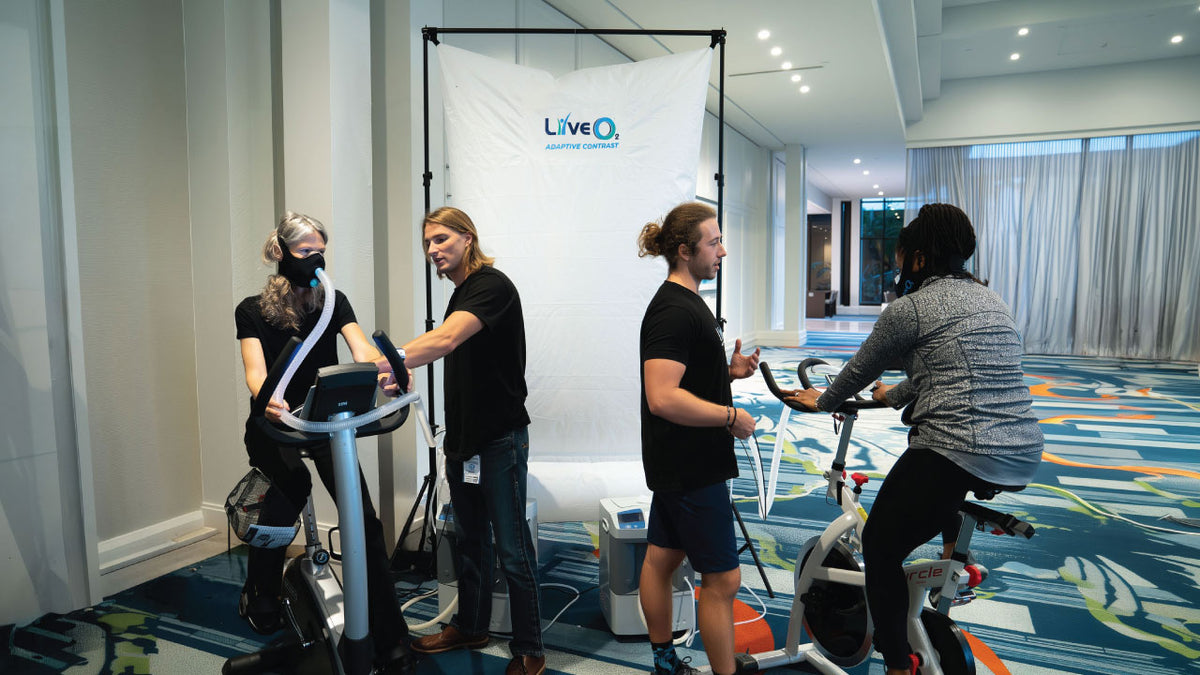 LiveO2 Dual-User Adaptive Contrast EWOT system with two users training on exercise bikes while staff assist in a professional facility setting