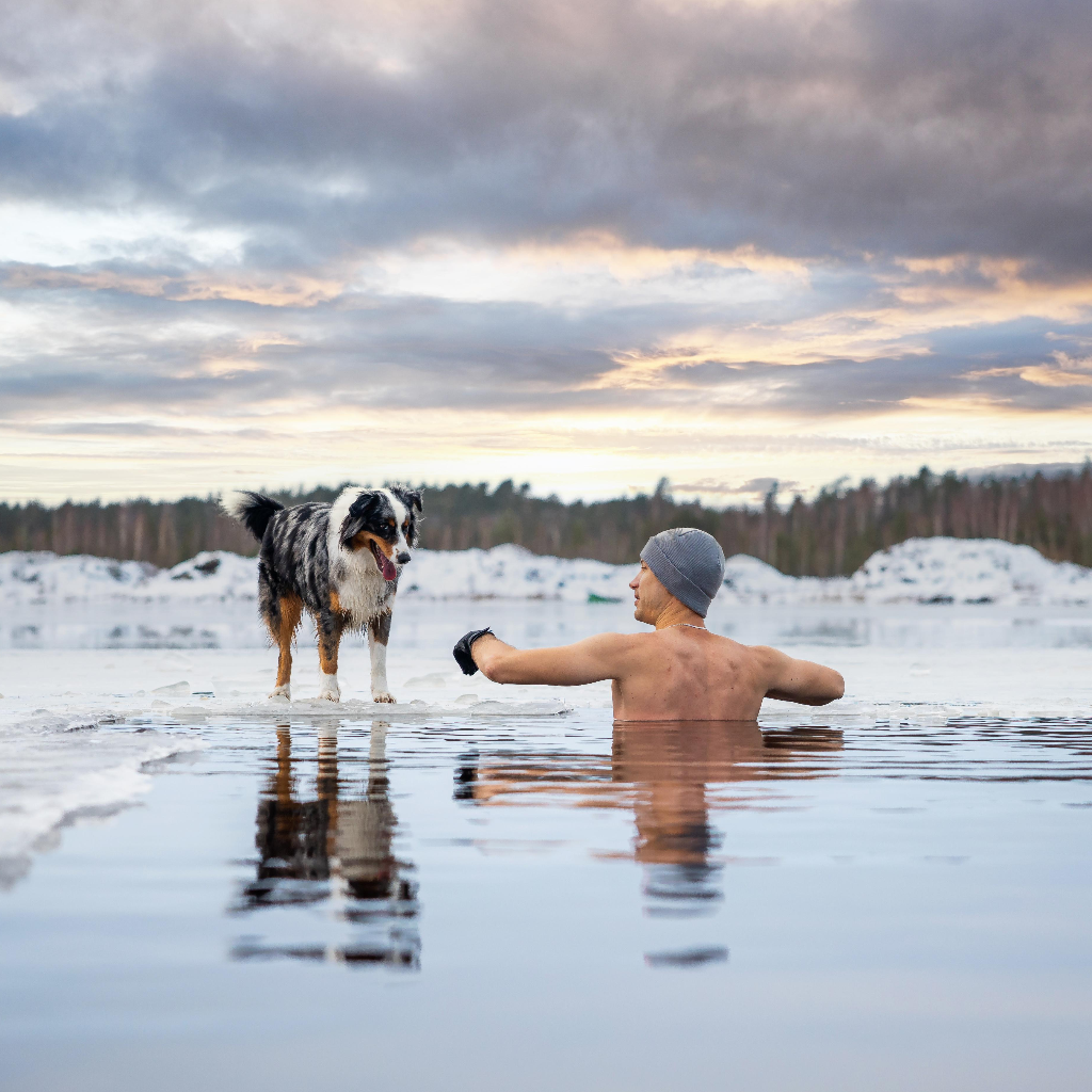 Man practicing cold water immersion in a frozen lake during winter while his dog stands on the ice nearby, illustrating cold exposure, resilience, and outdoor recovery.
